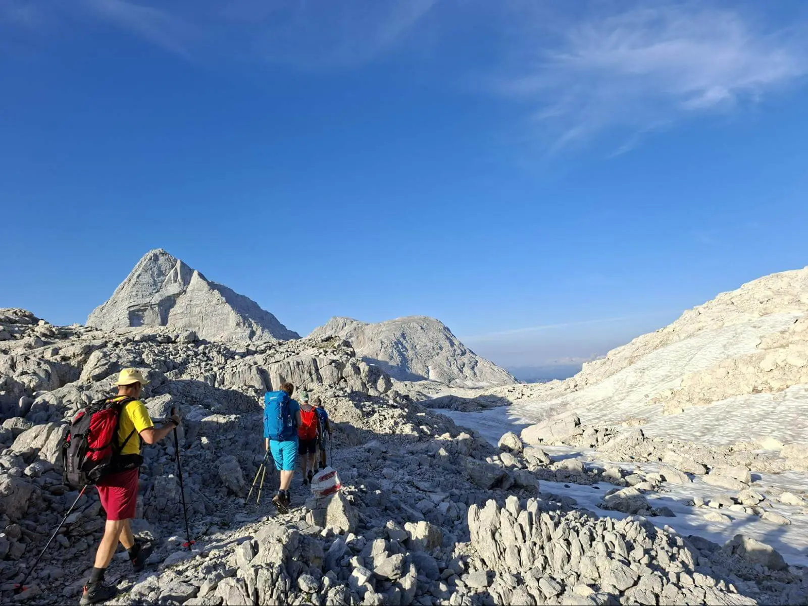 Mehrere Wandernde durchqueren eine helle Karstlandschaft mit Schneefeldern. Markante Gipfel ragen unter blauem Himmel empor. | © DAV Markt Schwaben/Alfred Quiel
