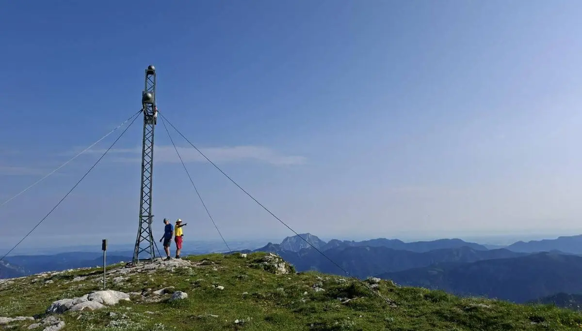 Zwei Wandernde stehen neben einem Gipfelkreuz auf dem Schönberg. Dahinter erstreckt sich eine weite Berglandschaft unter klarem Himmel. | © DAV Markt Schwaben/Alfred Quiel
