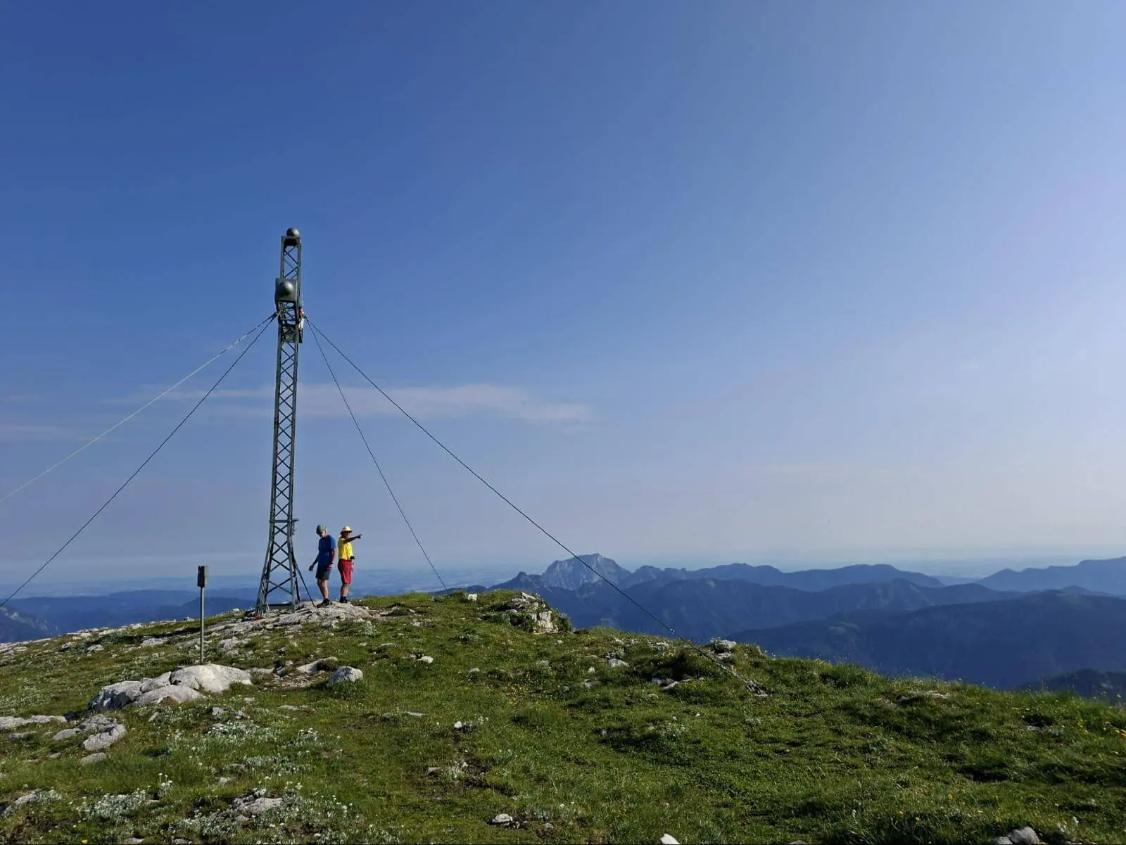 Zwei Wandernde stehen neben einem Gipfelkreuz auf dem Schönberg. Dahinter erstreckt sich eine weite Berglandschaft unter klarem Himmel. | © DAV Markt Schwaben/Alfred Quiel