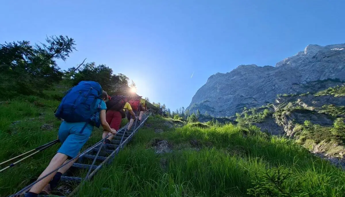 Mehrere Wandernde steigen über eine Metallleiter einen steilen Bergpfad hinauf. Im Hintergrund ragen Felswände unter klarem Himmel auf. | © DAV Markt Schwaben/Alfred Quiel