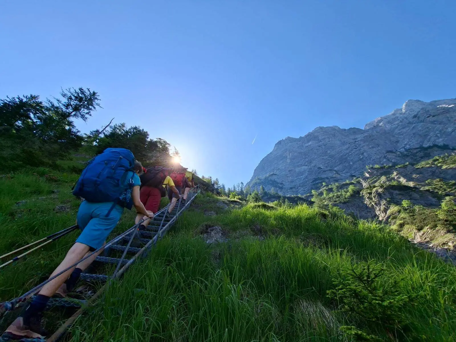 Mehrere Wandernde steigen über eine Metallleiter einen steilen Bergpfad hinauf. Im Hintergrund ragen Felswände unter klarem Himmel auf. | © DAV Markt Schwaben/Alfred Quiel