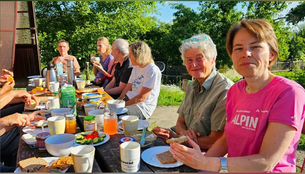 Tourenteilnehmende sitzen an einem reich gedeckten Tisch vor der Hütte. Brot, Aufstriche und Getränke stehen im Sonnenschein bereit. | © DAV Markt Schwaben/Lutz Gründel