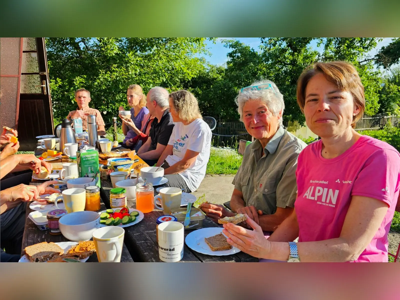 Tourenteilnehmende sitzen an einem reich gedeckten Tisch vor der Hütte. Brot, Aufstriche und Getränke stehen im Sonnenschein bereit. | © DAV Markt Schwaben/Lutz Gründel