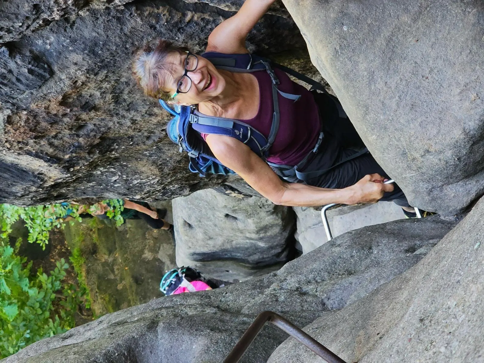 Eine Wandernde steigt auf Eisenstufen durch eine enge Felsspalte zwischen großen Sandsteinblöcken; weitere Personen folgen darunter. | © DAV Markt Schwaben/Lutz Gründel