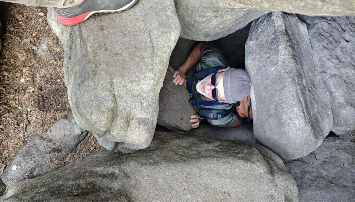 Eine Person zwängt sich mit Rucksack durch eine schmale Öffnung zwischen großen Sandsteinblöcken; Wanderschuhe sind am oberen Rand sichtbar. | © DAV Markt Schwaben/Erwin&Ursula Matzinger