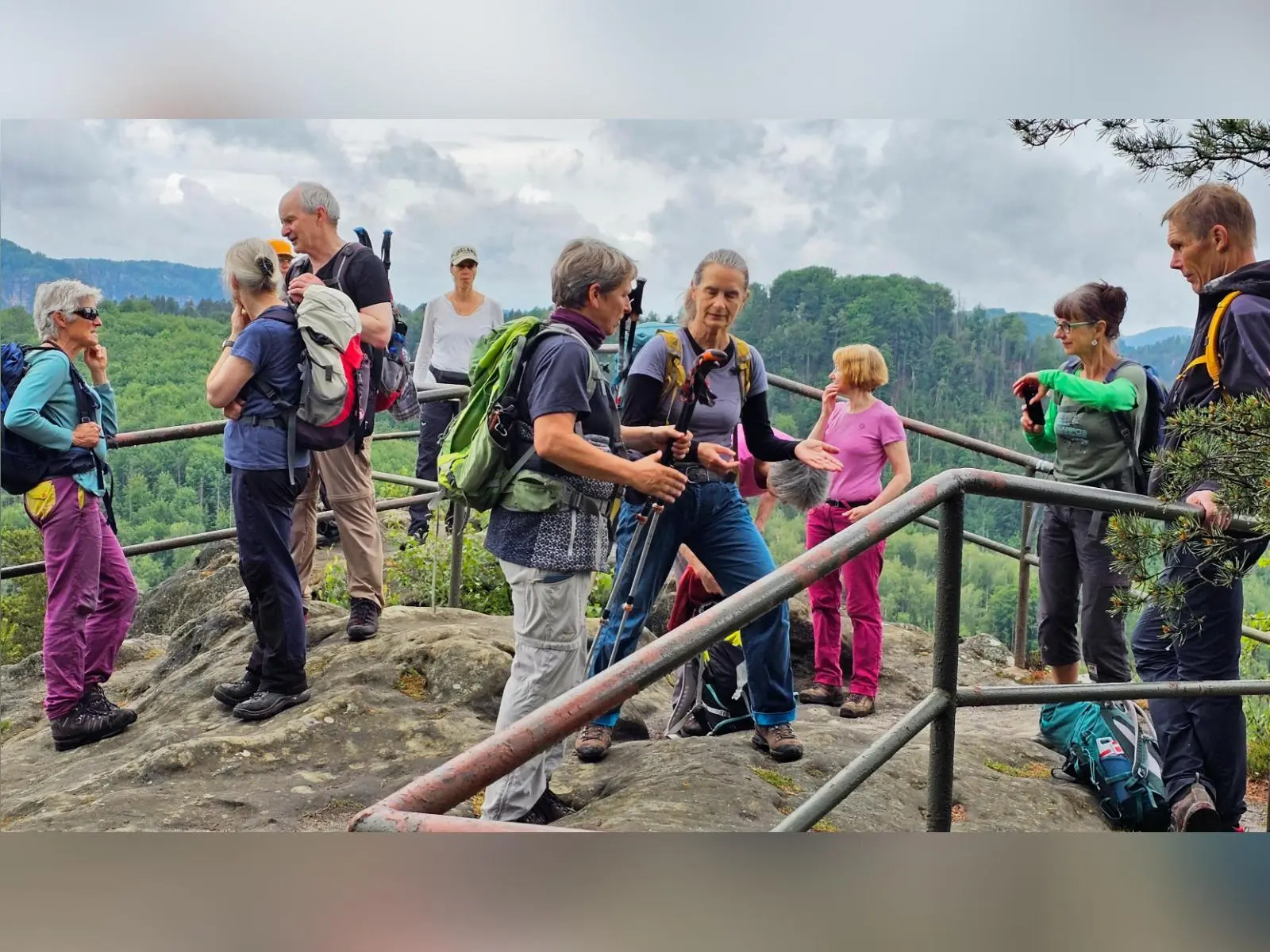 Eine Wandergruppe steht auf einem felsigen Aussichtspunkt mit Geländer. Im Hintergrund erstrecken sich bewaldete Hügel und Sandsteinfelsen der Sächsischen Schweiz. | © DAV Markt Schwaben/Lutz Gründel