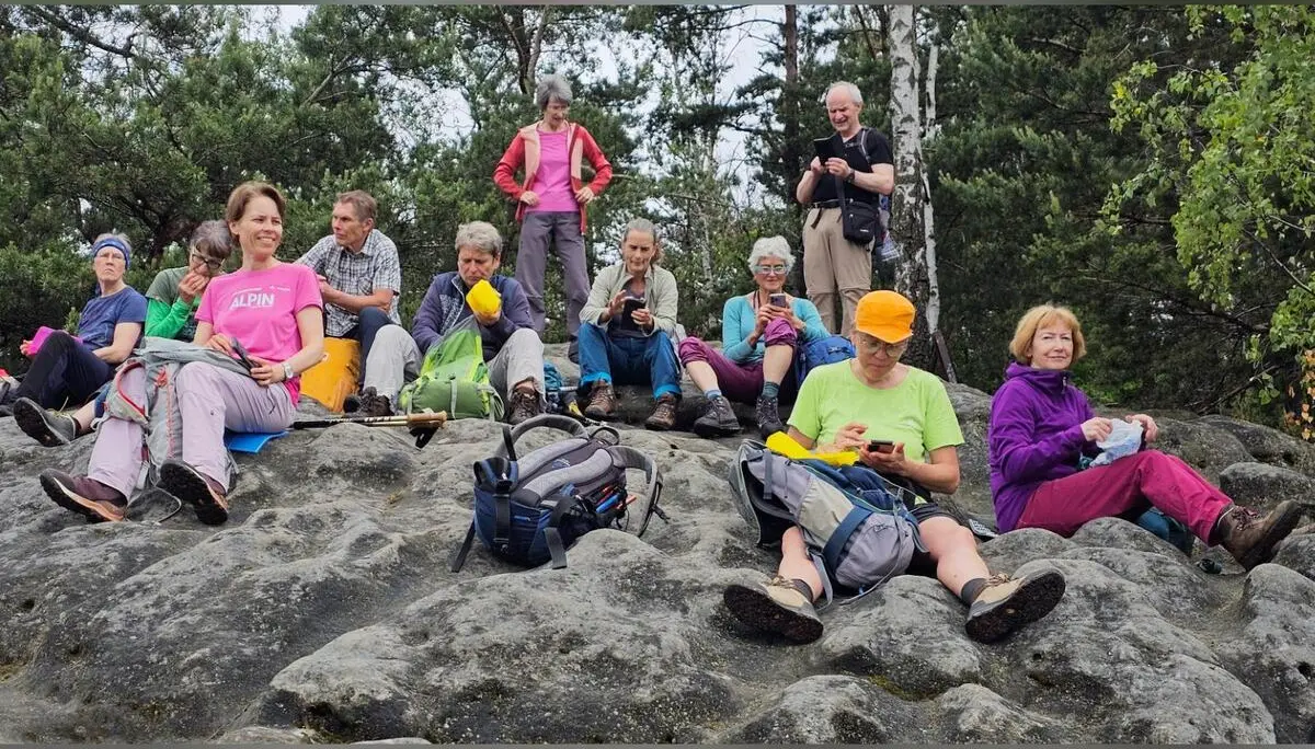 Mehrere Tourenteilnehmende sitzen mit Rucksäcken auf einer Sandsteinfläche während einer Pause. Dahinter stehen Kiefern und Birken am Rand des Aussichtsfelsens. | © DAV Markt Schwaben/Lutz Gründel
