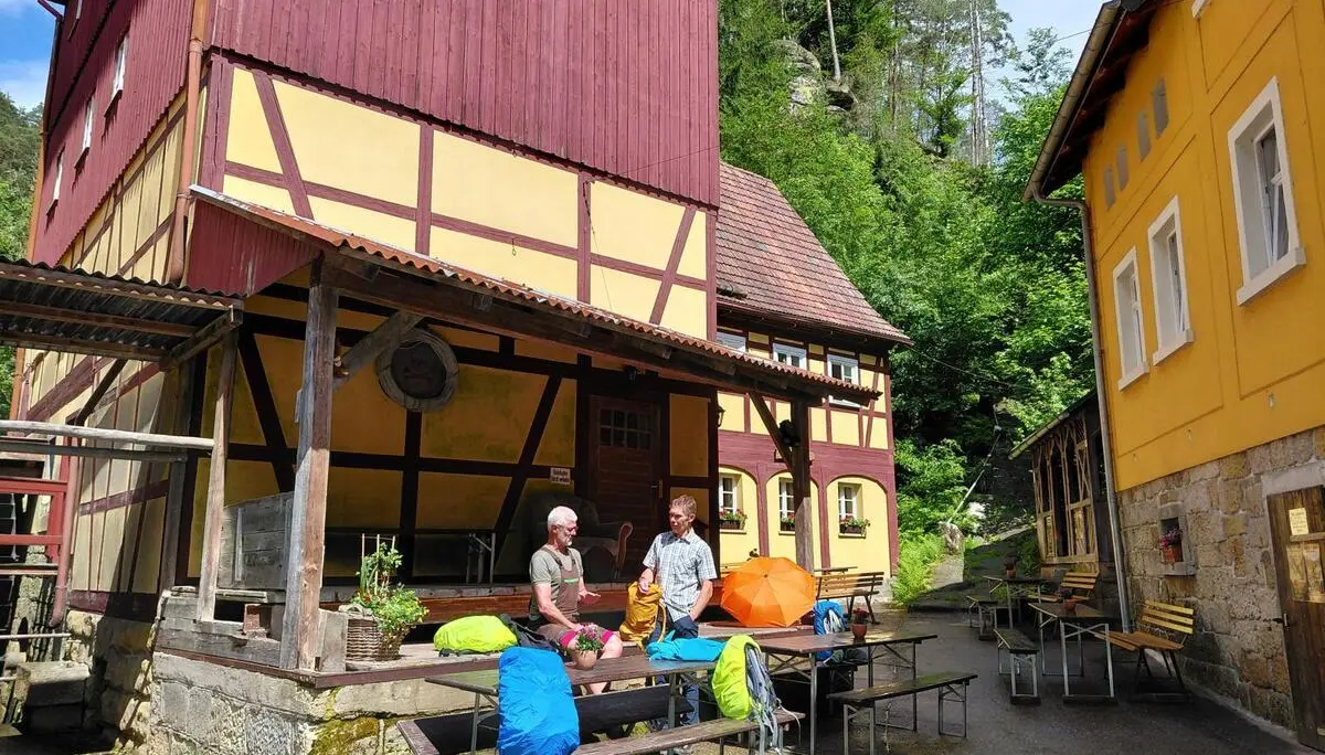 Zwei Tourenteilnehmende stehen im Innenhof der Dessauer Hütte zwischen Fachwerkgebäuden. Vor ihnen liegen Rucksäcke und Biertischgarnituren im Sonnenschein. | © DAV Markt Schwaben/Sabine Hainz