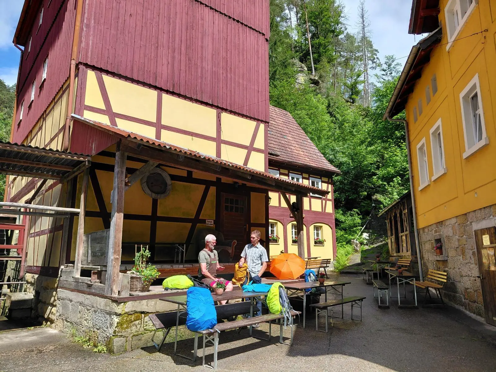 Zwei Tourenteilnehmende stehen im Innenhof der Dessauer Hütte zwischen Fachwerkgebäuden. Vor ihnen liegen Rucksäcke und Biertischgarnituren im Sonnenschein. | © DAV Markt Schwaben/Sabine Hainz