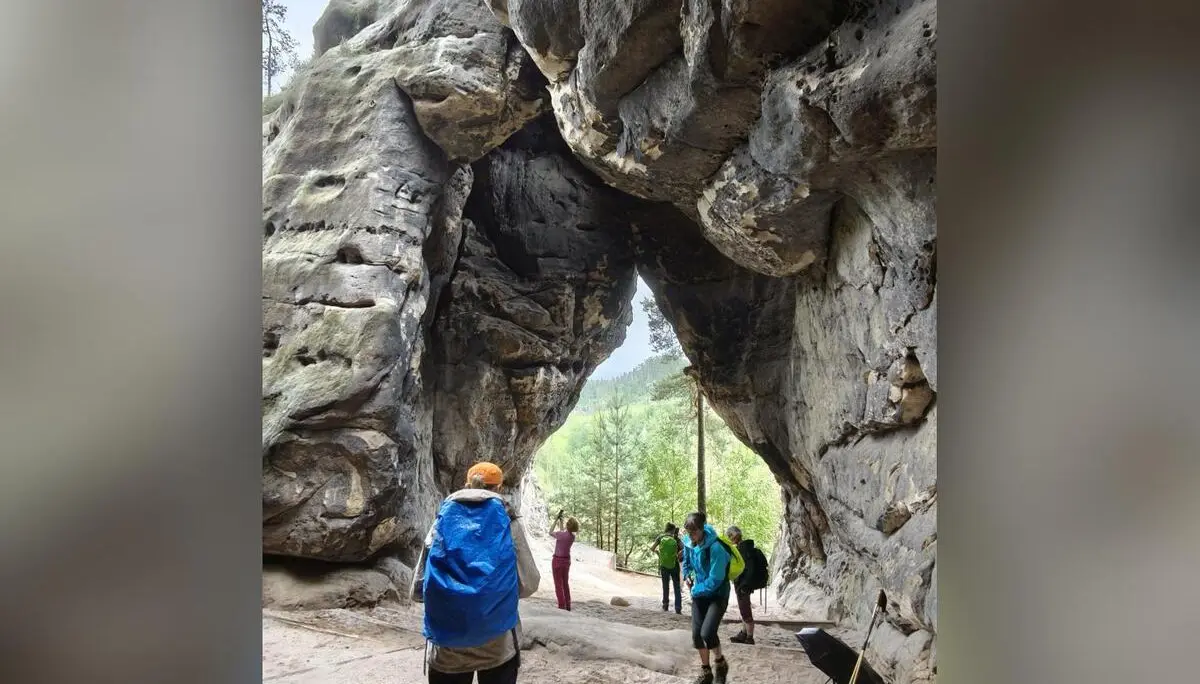 Mehrere Wandernde stehen unter einem imposanten natürlichen Felsbogen aus Sandstein. Durch die Öffnung fällt Licht auf den Weg und die bewaldete Landschaft dahinter. | © DAV Markt Schwaben/Erwin&Ursula Matzinger
