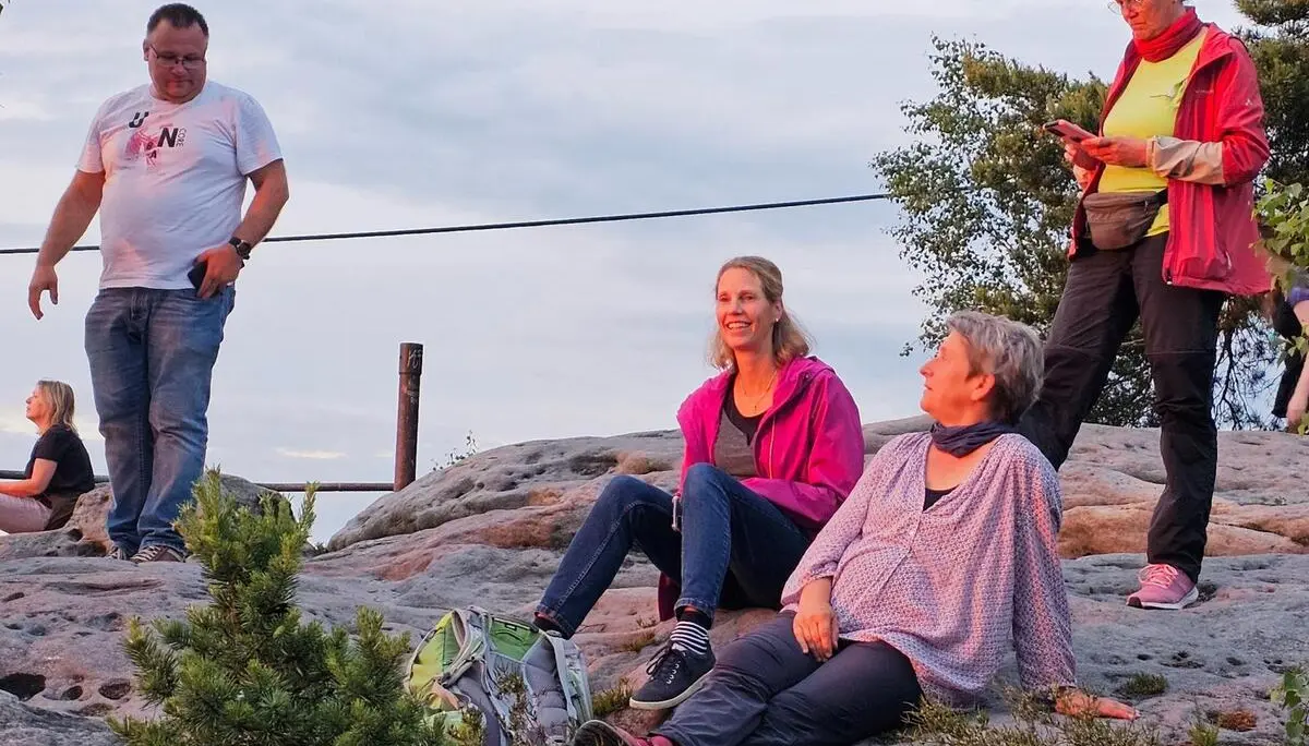 Mehrere Tourenteilnehmende sitzen und stehen auf einer Sandsteinfläche beim Sonnenuntergang. Warmes Abendlicht beleuchtet Felsen, Rucksäcke und die Aussichtskanzel. | © DAV Markt Schwaben/Lutz Gründel