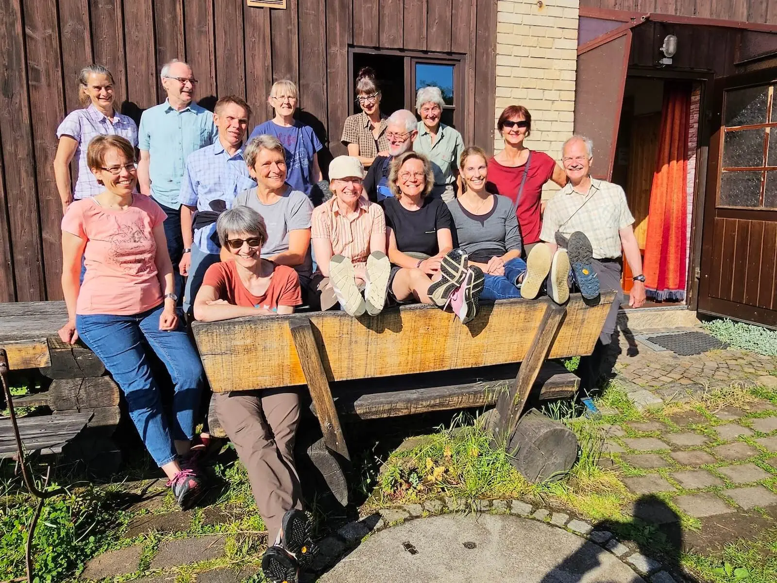 Die Tourengruppe versammelt sich für ein Gruppenfoto vor einem Gebäude der Dessauer Hütte. Einige Personen sitzen auf einer großen Holzbank, andere stehen dahinter im Sonnenlicht. | © DAV Markt Schwaben/Lutz Gründel