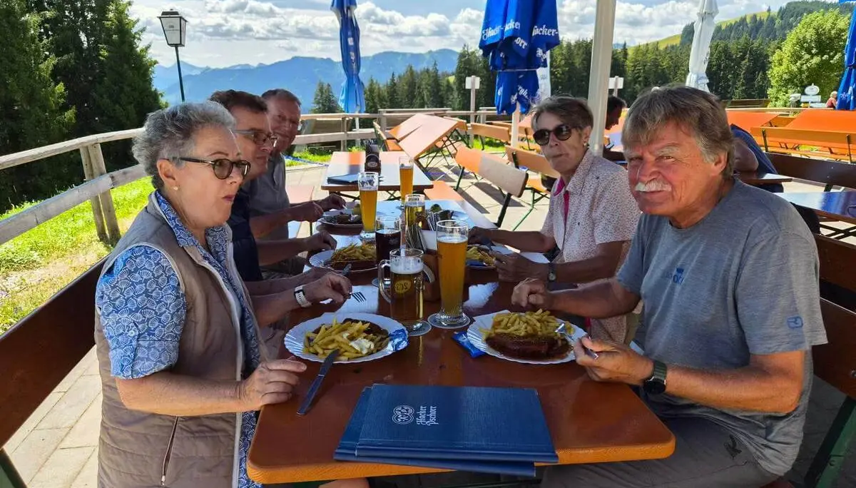 Fünf Personen sitzen auf der Terrasse eines Berggasthauses beim Mittagessen; im Hintergrund sind Berge und Wald zu sehen. | © DAV Markt Schwaben / Hubert Inhofer