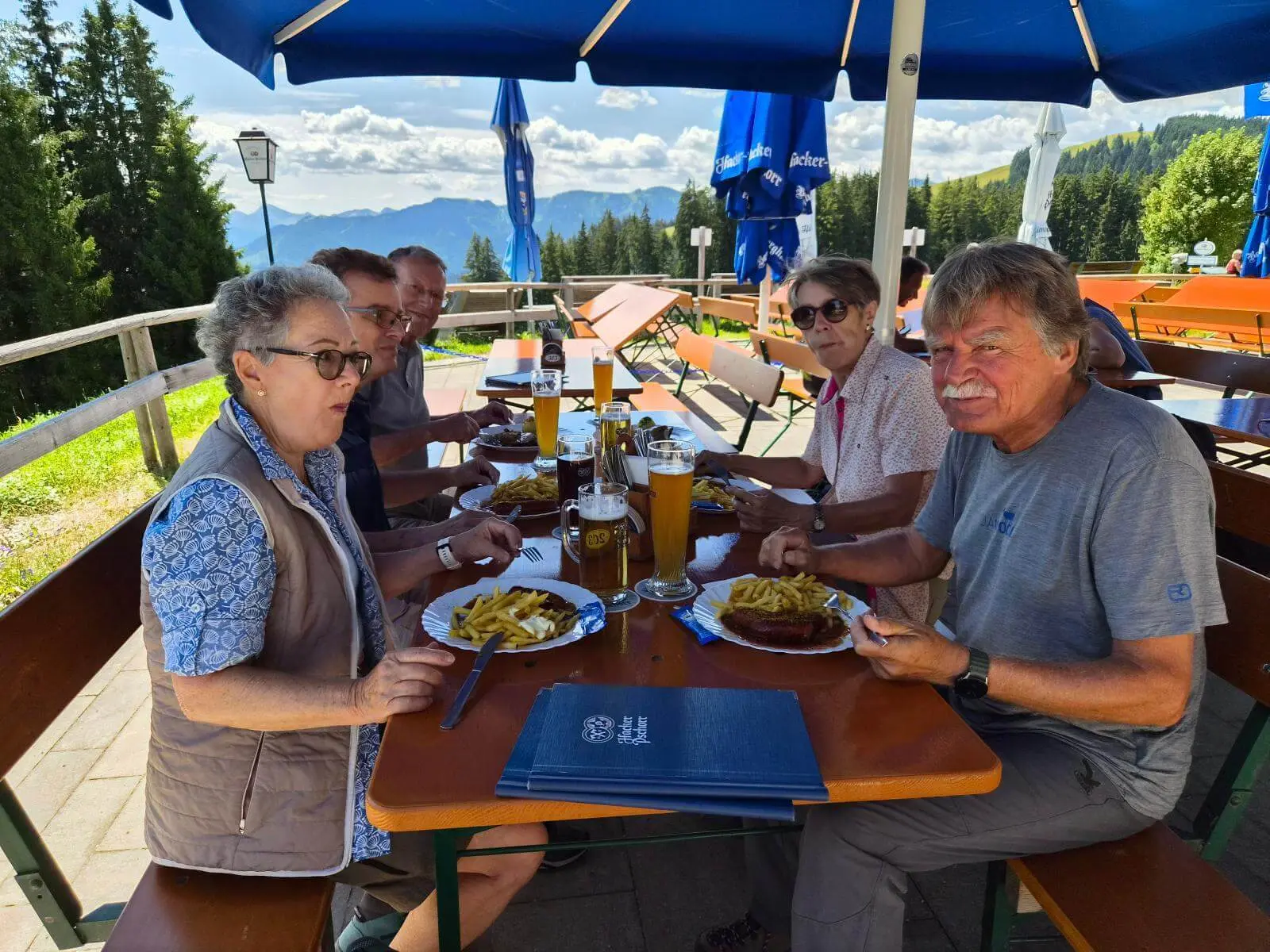 Fünf Personen sitzen auf der Terrasse eines Berggasthauses beim Mittagessen; im Hintergrund sind Berge und Wald zu sehen. | © DAV Markt Schwaben / Hubert Inhofer