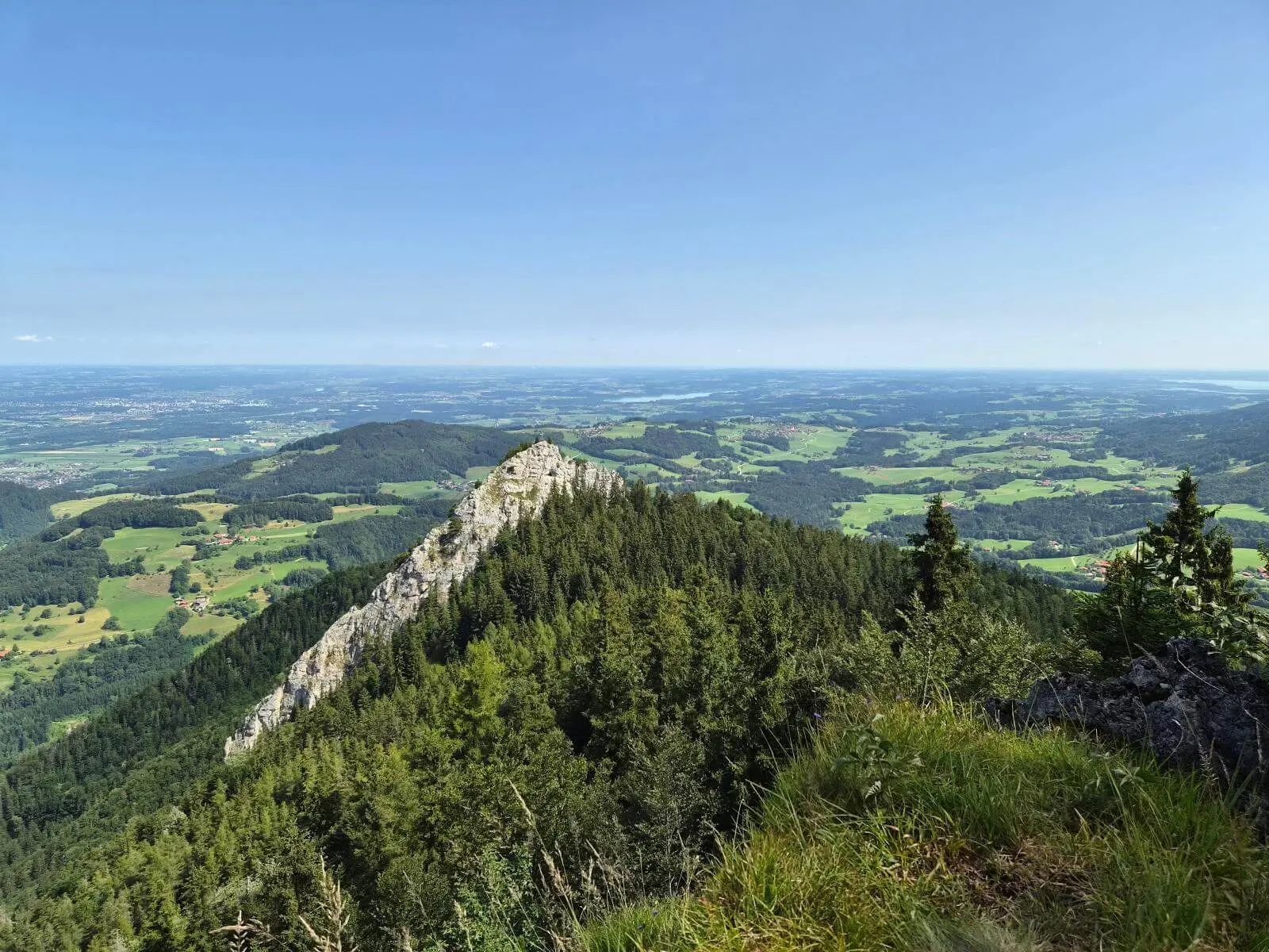 Aussicht von einer Bergwiese auf einen bewaldeten Felsrücken und das weite Alpenvorland. Felder, Wälder und Ortschaften prägen die Landschaft. | © DAV Markt Schwaben / Hubert Inhofer