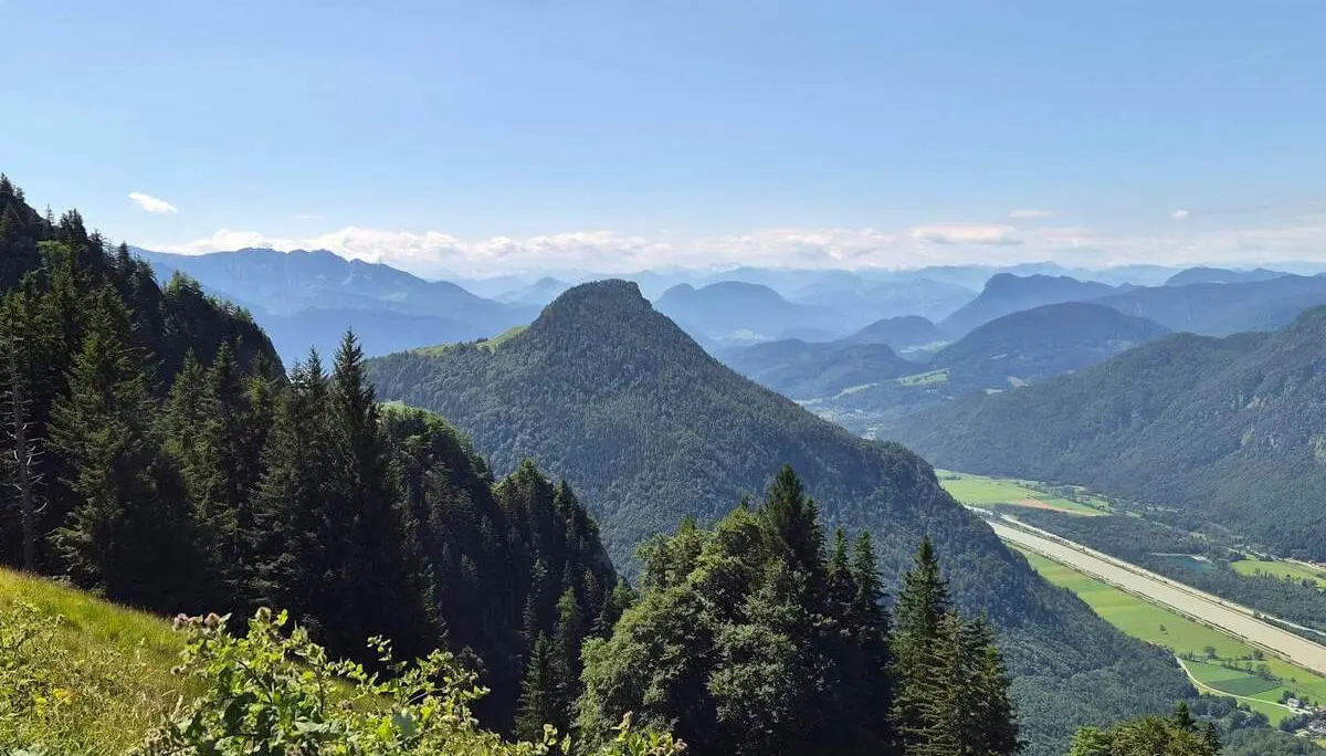 Weite Aussicht über bewaldete Berge und das Inntal mit Feldern im Talboden. Im Vordergrund liegen eine Bergwiese und Nadelbäume.  | © DAV Markt Schwaben / Hubert Inhofer