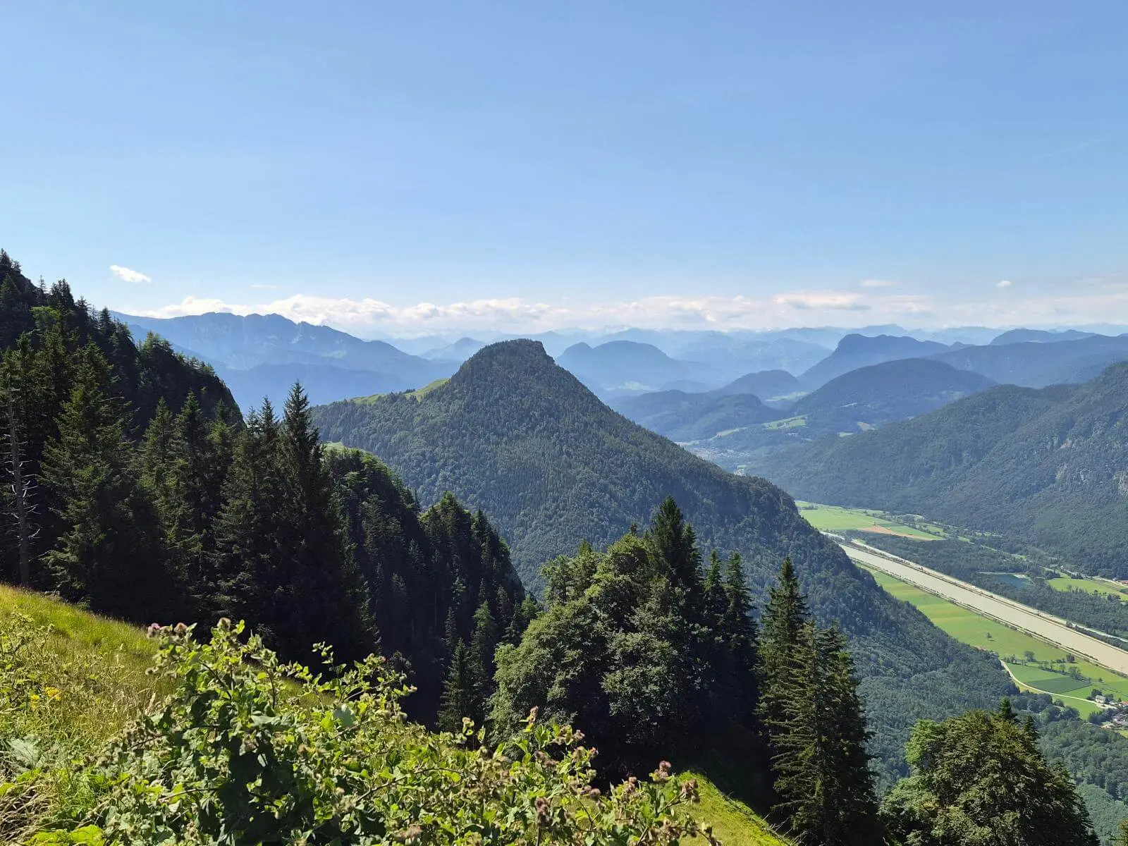 Weite Aussicht über bewaldete Berge und das Inntal mit Feldern im Talboden. Im Vordergrund liegen eine Bergwiese und Nadelbäume.  | © DAV Markt Schwaben / Hubert Inhofer
