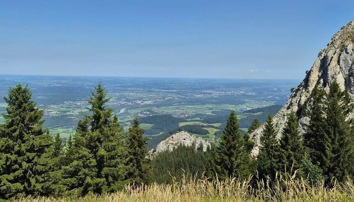 Aussicht über eine Bergwiese und Nadelbäume ins Inntal. Rechts ragt eine steile Felswand auf, dahinter liegen Felder und Ortschaften. | © DAV Markt Schwaben / Hubert Inhofer