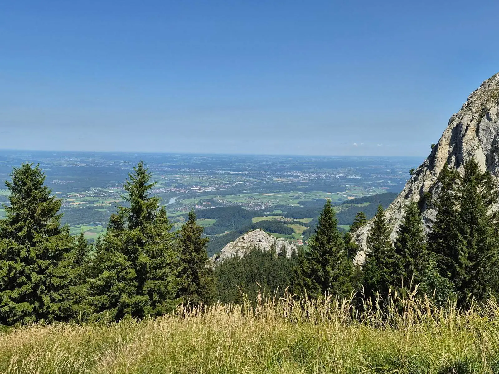 Aussicht über eine Bergwiese und Nadelbäume ins Inntal. Rechts ragt eine steile Felswand auf, dahinter liegen Felder und Ortschaften. | © DAV Markt Schwaben / Hubert Inhofer
