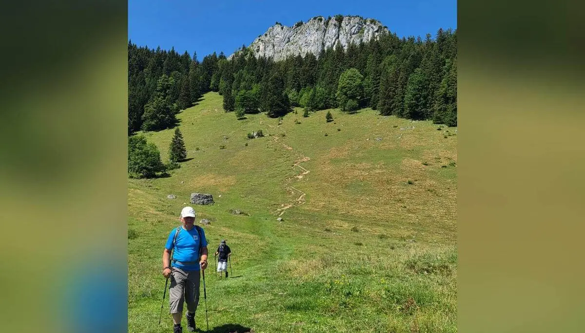 Zwei Wandernde mit Trekkingstöcken gehen über eine offene Bergwiese. Darüber erhebt sich eine markante Felswand am bewaldeten Berghang. | © DAV Markt Schwaben / Hubert Inhofer