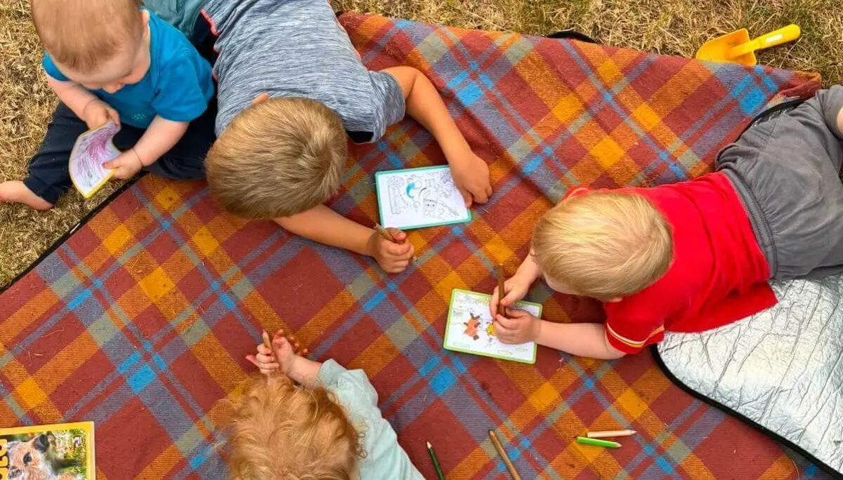 Vier Kinder sitzen und liegen auf einer karierten Decke im Gras und malen mit Buntstiften in kleinen Malbüchern. | © DAV Markt Schwaben / Lara Wolff