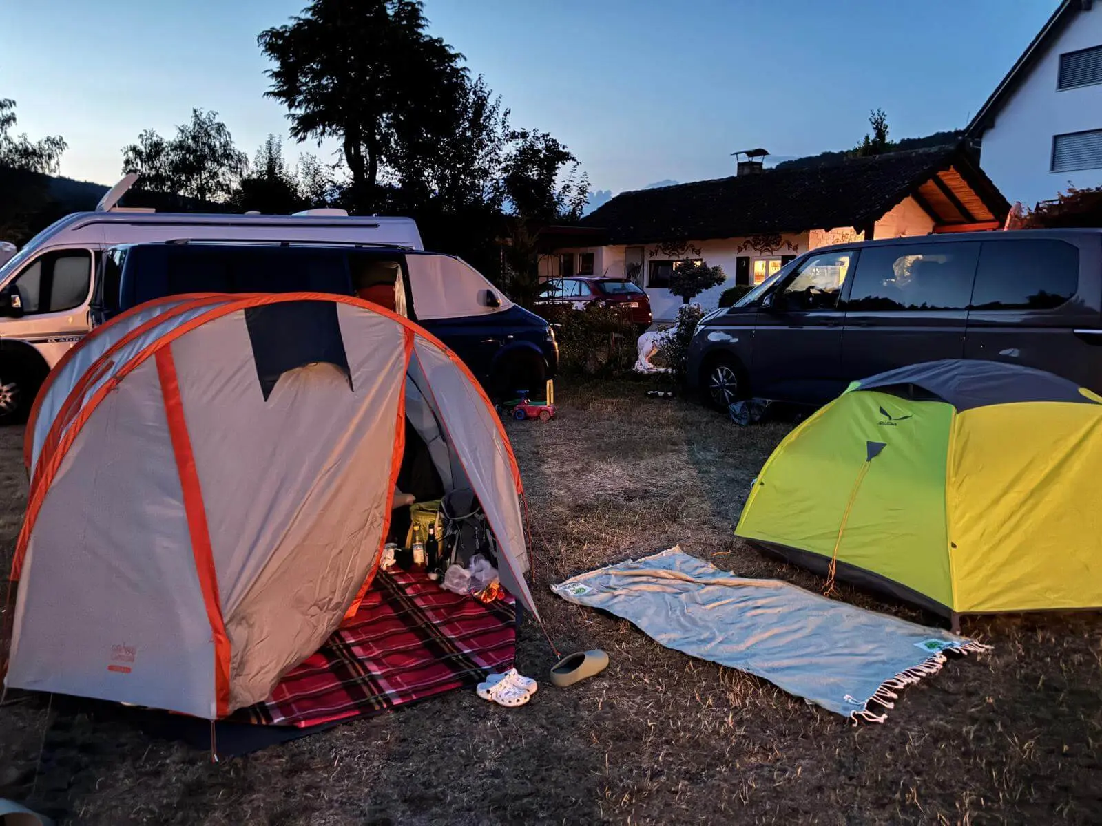 Zwei Zelte stehen zwischen Campingbussen auf einem trockenen Zeltplatz. Die Szene ist in der Abenddämmerung beleuchtet. | © DAV Markt Schwaben / Lara Wolff