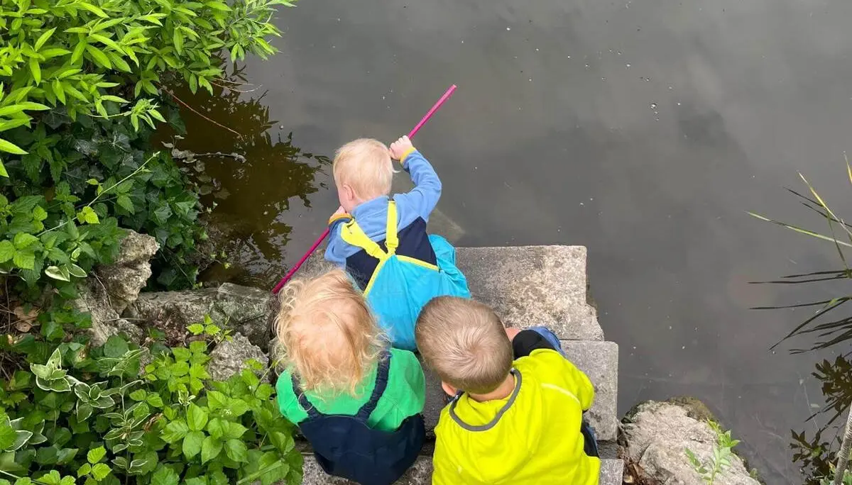 Drei Kinder sitzen auf Steinstufen an einem Teich. Ein Kind hält einen Stock ins Wasser, um die Oberfläche zu erkunden. | © DAV Markt Schwaben / Lara Wolff
