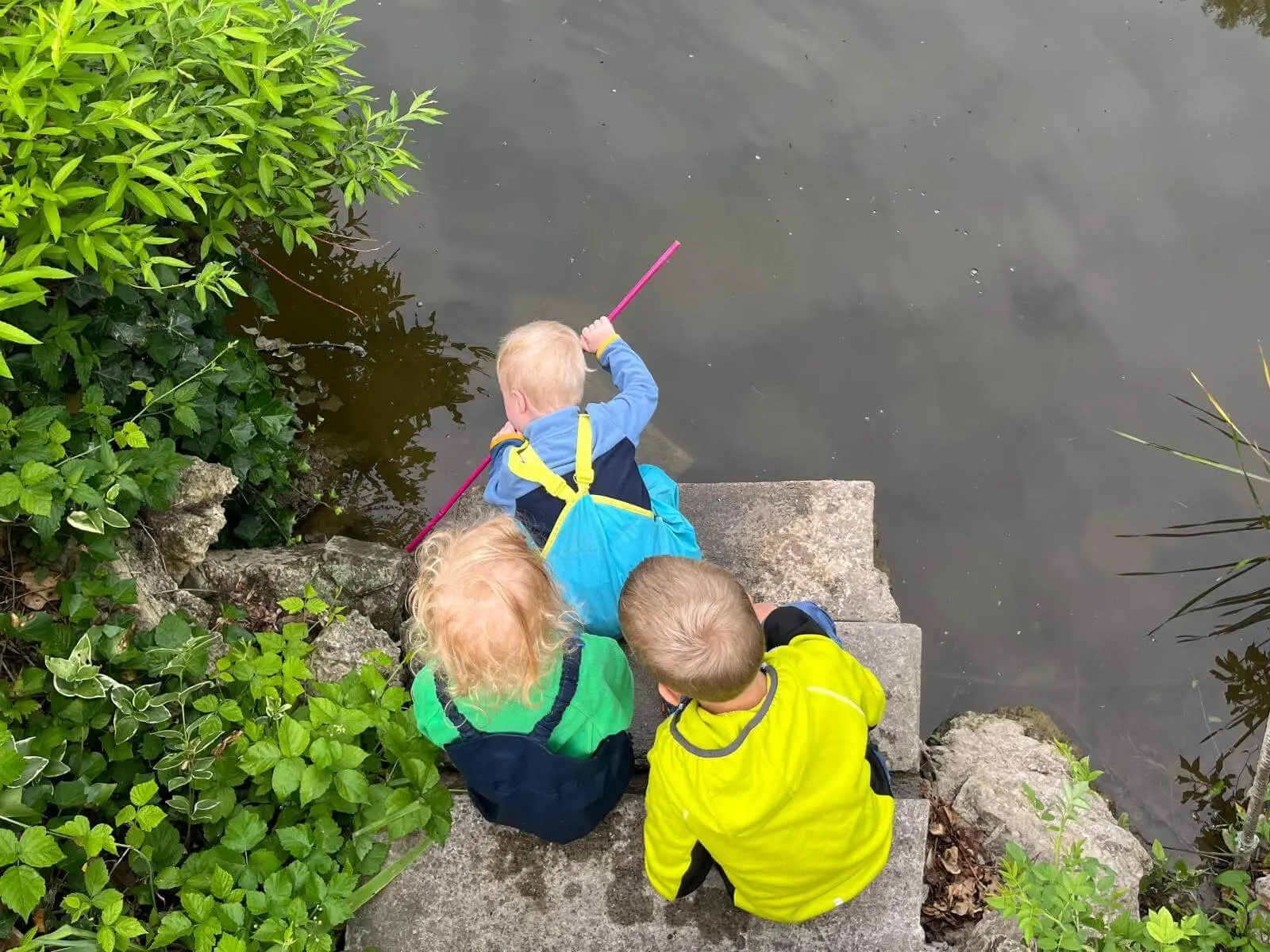 Drei Kinder sitzen auf Steinstufen an einem Teich. Ein Kind hält einen Stock ins Wasser, um die Oberfläche zu erkunden. | © DAV Markt Schwaben / Lara Wolff