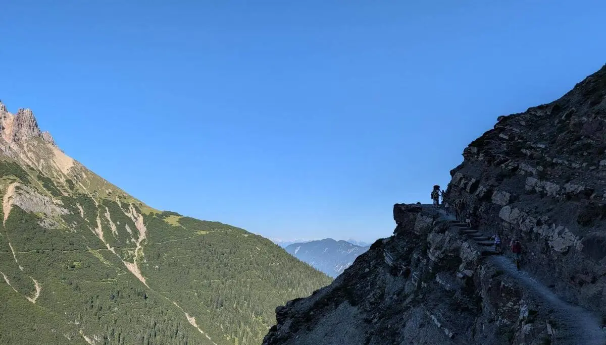 Wandernde bewegen sich auf einem schmalen, in den Fels gehauenen Bergweg über einem steilen Hang. Im Hintergrund liegen bewaldete Bergrücken und Gipfel. | © DAV Markt Schwaben / Silke Titze