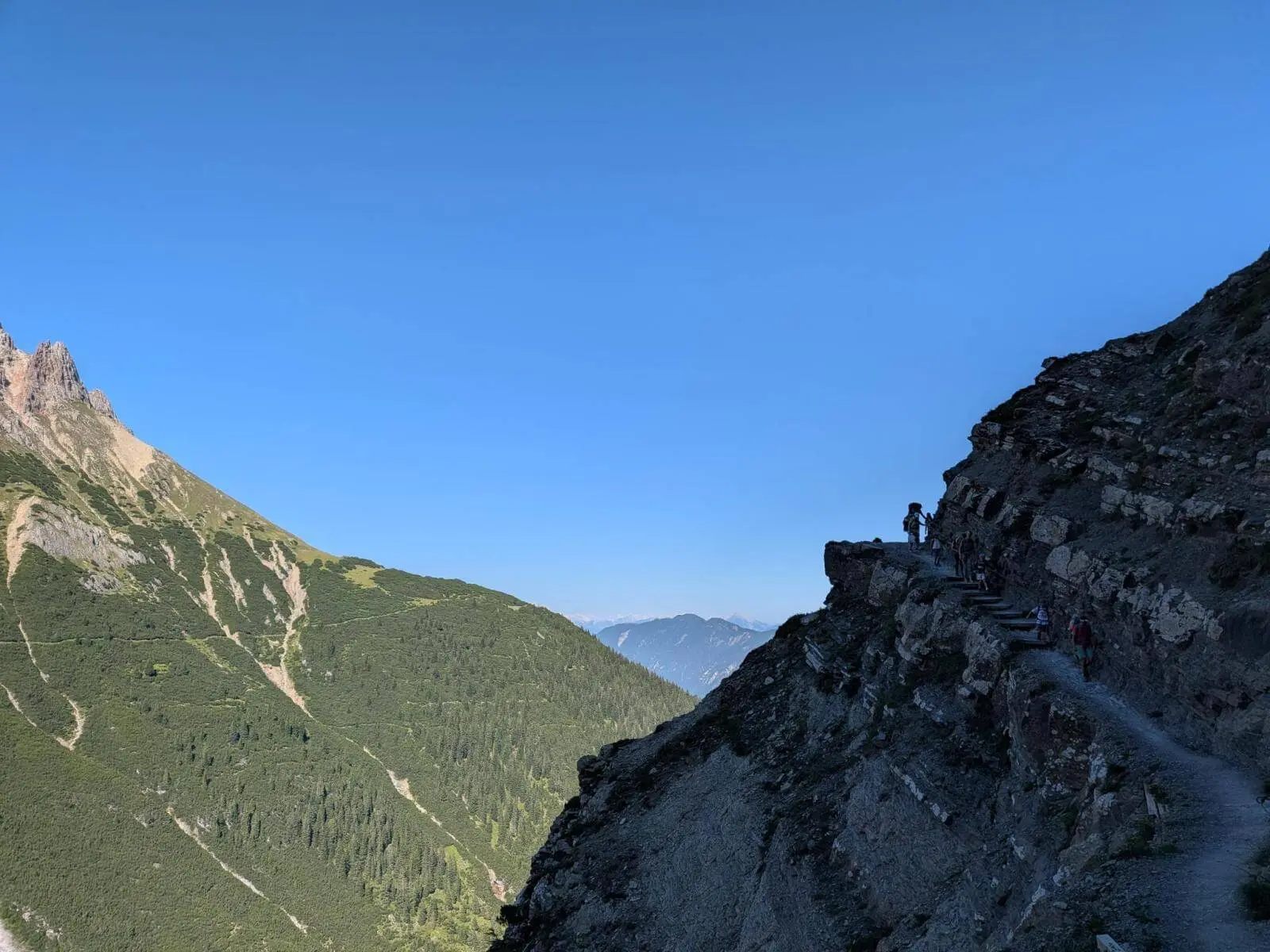 Wandernde bewegen sich auf einem schmalen, in den Fels gehauenen Bergweg über einem steilen Hang. Im Hintergrund liegen bewaldete Bergrücken und Gipfel. | © DAV Markt Schwaben / Silke Titze