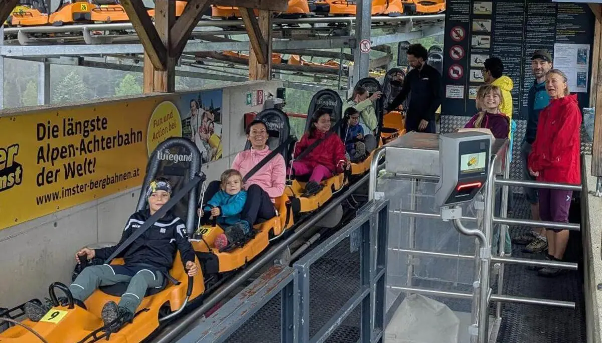 Kinder und Erwachsene sitzen angeschnallt in Alpine-Coaster-Fahrzeugen an der Talstation. Die Bahn steht zur Abfahrt bereit. | © DAV Markt Schwaben / Silke Titze