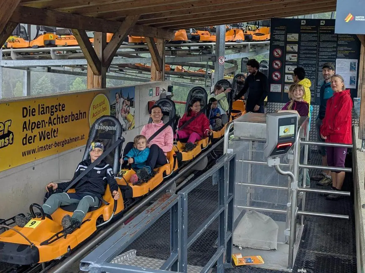 Kinder und Erwachsene sitzen angeschnallt in Alpine-Coaster-Fahrzeugen an der Talstation. Die Bahn steht zur Abfahrt bereit. | © DAV Markt Schwaben / Silke Titze
