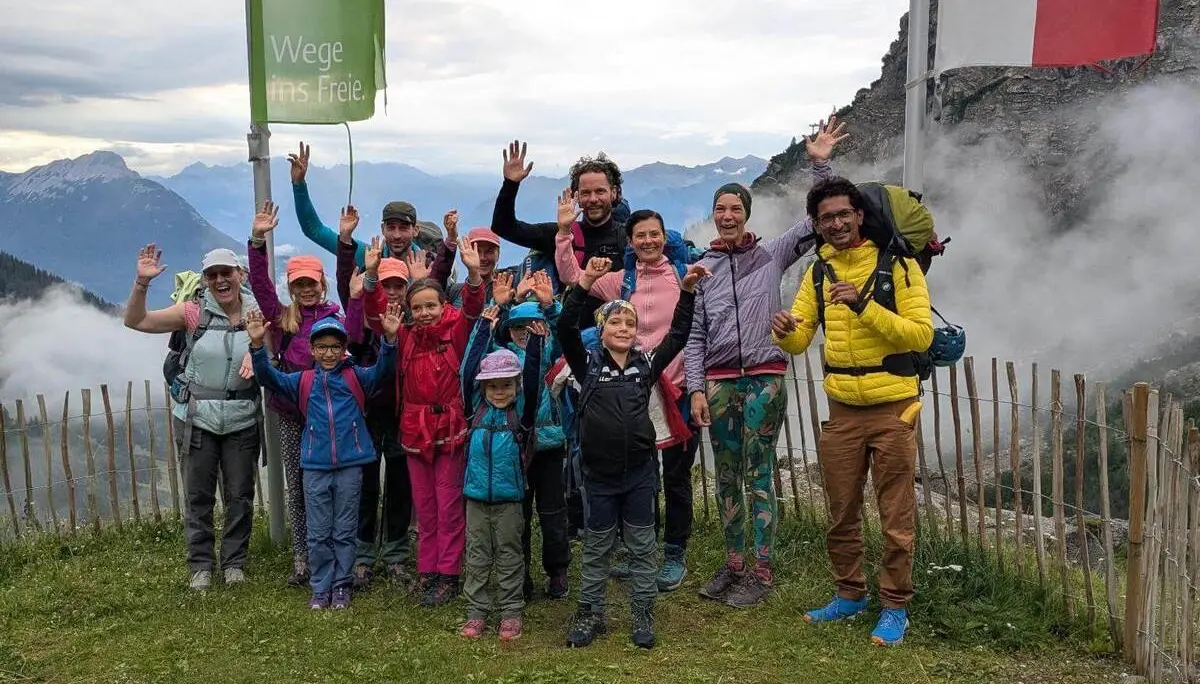 Kinder und Erwachsene stehen für ein Gruppenfoto an einer Bergstation und heben die Hände. Nebelschwaden ziehen vor den Bergen im Hintergrund vorbei. | © DAV Markt Schwaben / Silke Titze
