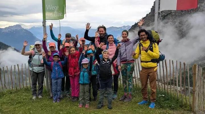 Kinder und Erwachsene stehen für ein Gruppenfoto an einer Bergstation und heben die Hände. Nebelschwaden ziehen vor den Bergen im Hintergrund vorbei. | © DAV Markt Schwaben / Silke Titze
