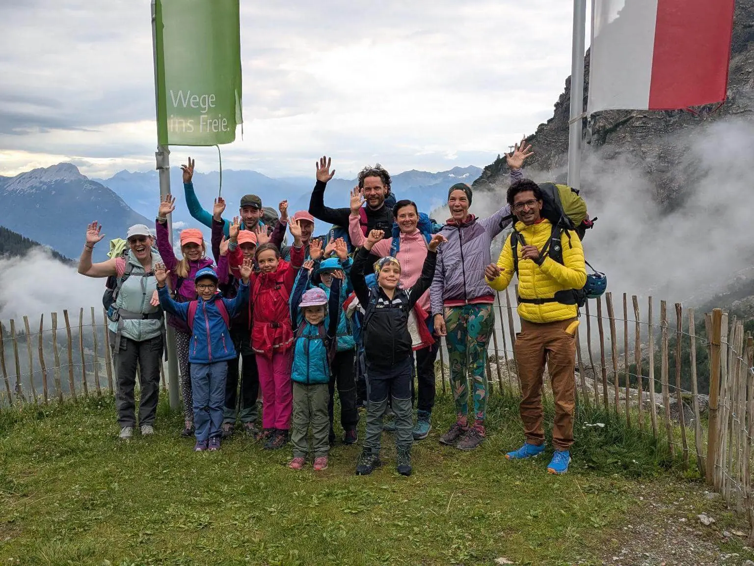 Kinder und Erwachsene stehen für ein Gruppenfoto an einer Bergstation und heben die Hände. Nebelschwaden ziehen vor den Bergen im Hintergrund vorbei. | © DAV Markt Schwaben / Silke Titze