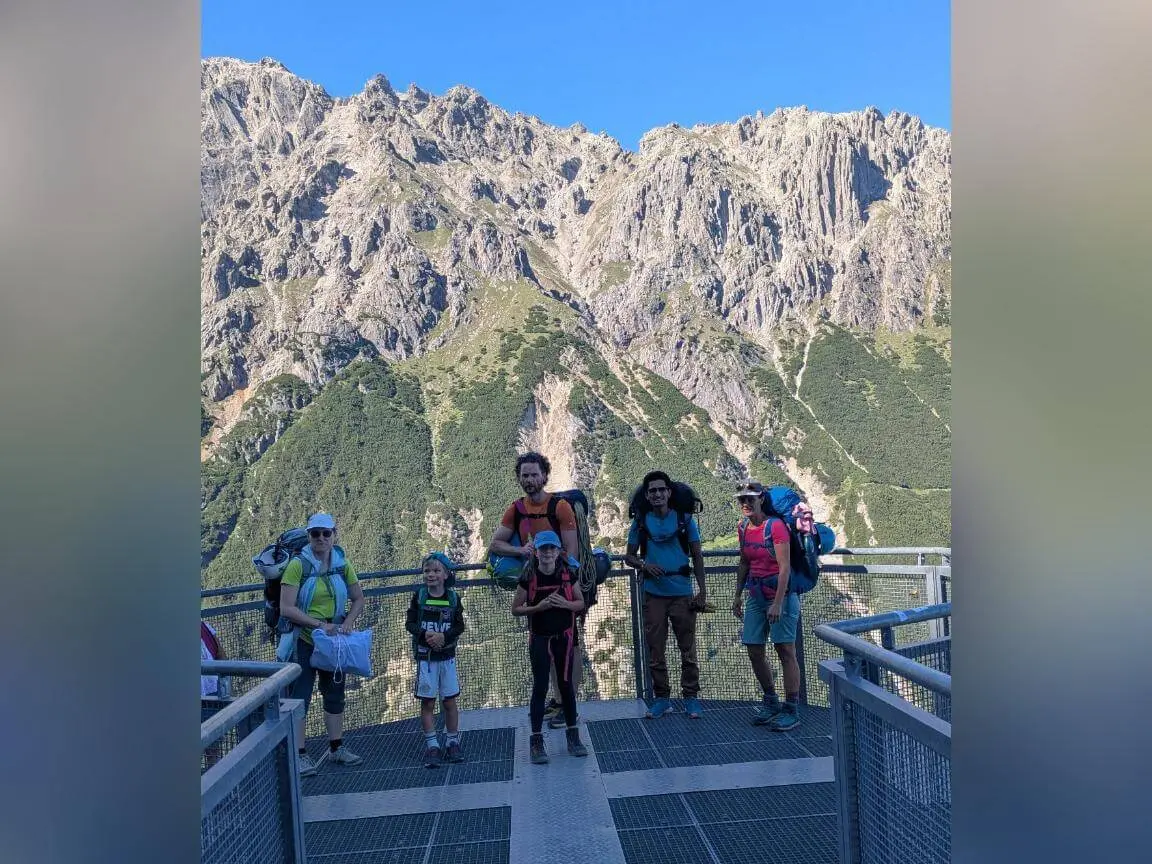 Fünf Wandernde mit Rucksäcken stehen auf einer Aussichtsplattform an der Bergstation. Dahinter ragen steile Felswände vor blauem Himmel auf. | © DAV Markt Schwaben / Silke Titze