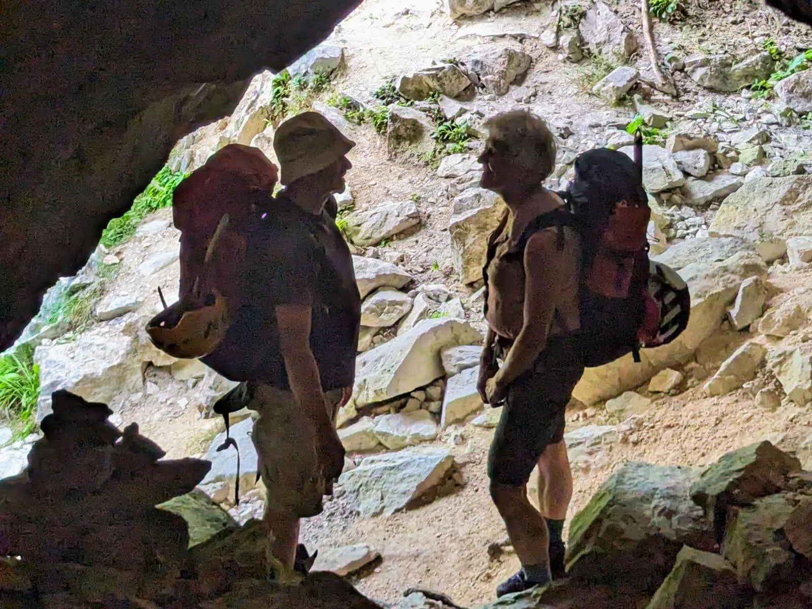 Zwei Wandernde mit großen Rucksäcken stehen im Eingang einer kleinen Höhle. Helles Tageslicht fällt auf den steinigen Bergpfad vor ihnen. | © DAV Markt Schwaben/Michael Huber