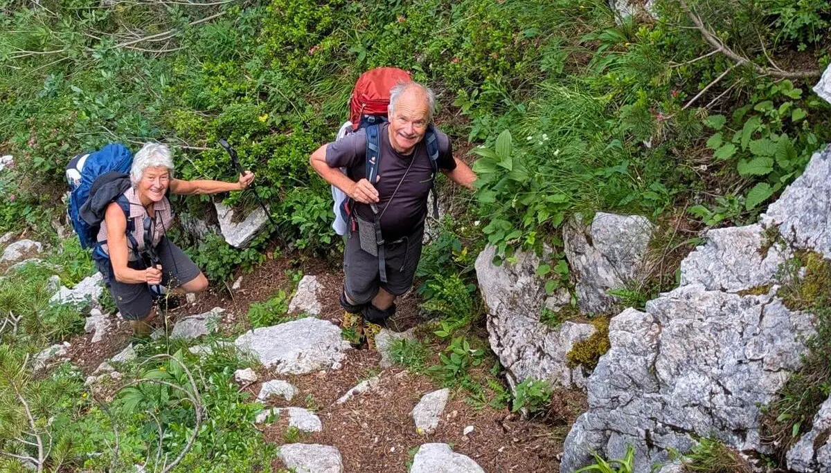 Zwei Wandernde mit großen Rucksäcken steigen auf einem steinigen Bergpfad zwischen Felsen, Sträuchern und alpiner Vegetation bergauf. | © DAV Markt Schwaben/Michael Huber