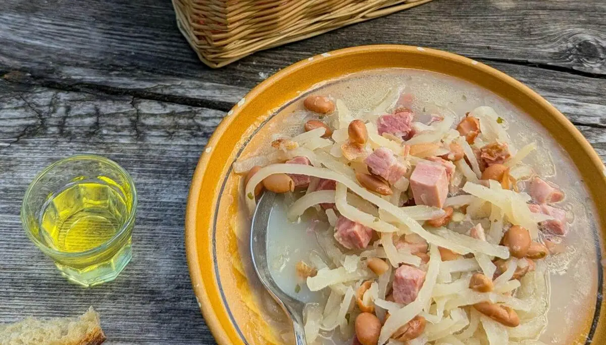 Eine Schüssel Jota mit Bohnen, Sauerkraut und Schinkenwürfeln steht auf einem Holztisch. Daneben liegen Brot und ein kleines Glas Getränk. | © DAV Markt Schwaben/Michael Huber