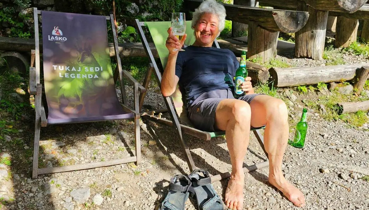 Eine Person sitzt barfuß in einem Liegestuhl vor einer Berghütte und hält ein Getränk. Daneben stehen weitere Flaschen und Wandersandalen. | © DAV Markt Schwaben/Lutz Gründel