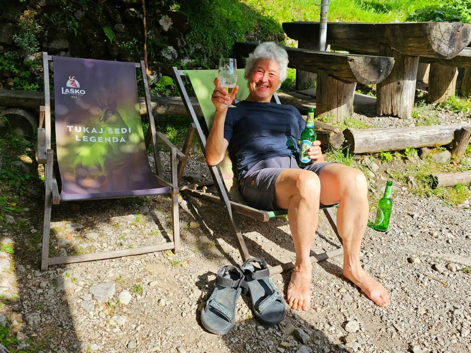 Eine Person sitzt barfuß in einem Liegestuhl vor einer Berghütte und hält ein Getränk. Daneben stehen weitere Flaschen und Wandersandalen. | © DAV Markt Schwaben/Lutz Gründel