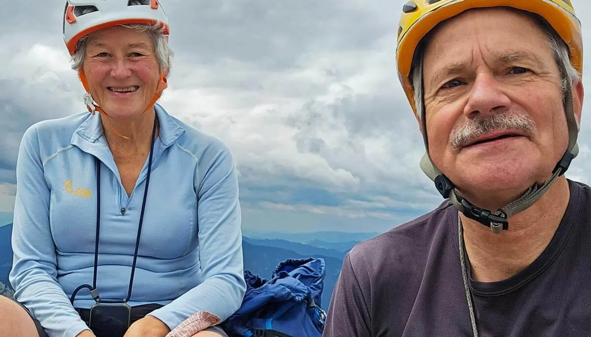 Zwei Personen mit Kletterhelmen sitzen auf einem Berggipfel vor wolkenverhangenem Himmel. Rucksäcke liegen neben ihnen am Boden. | © DAV Markt Schwaben/Lutz Gründel
