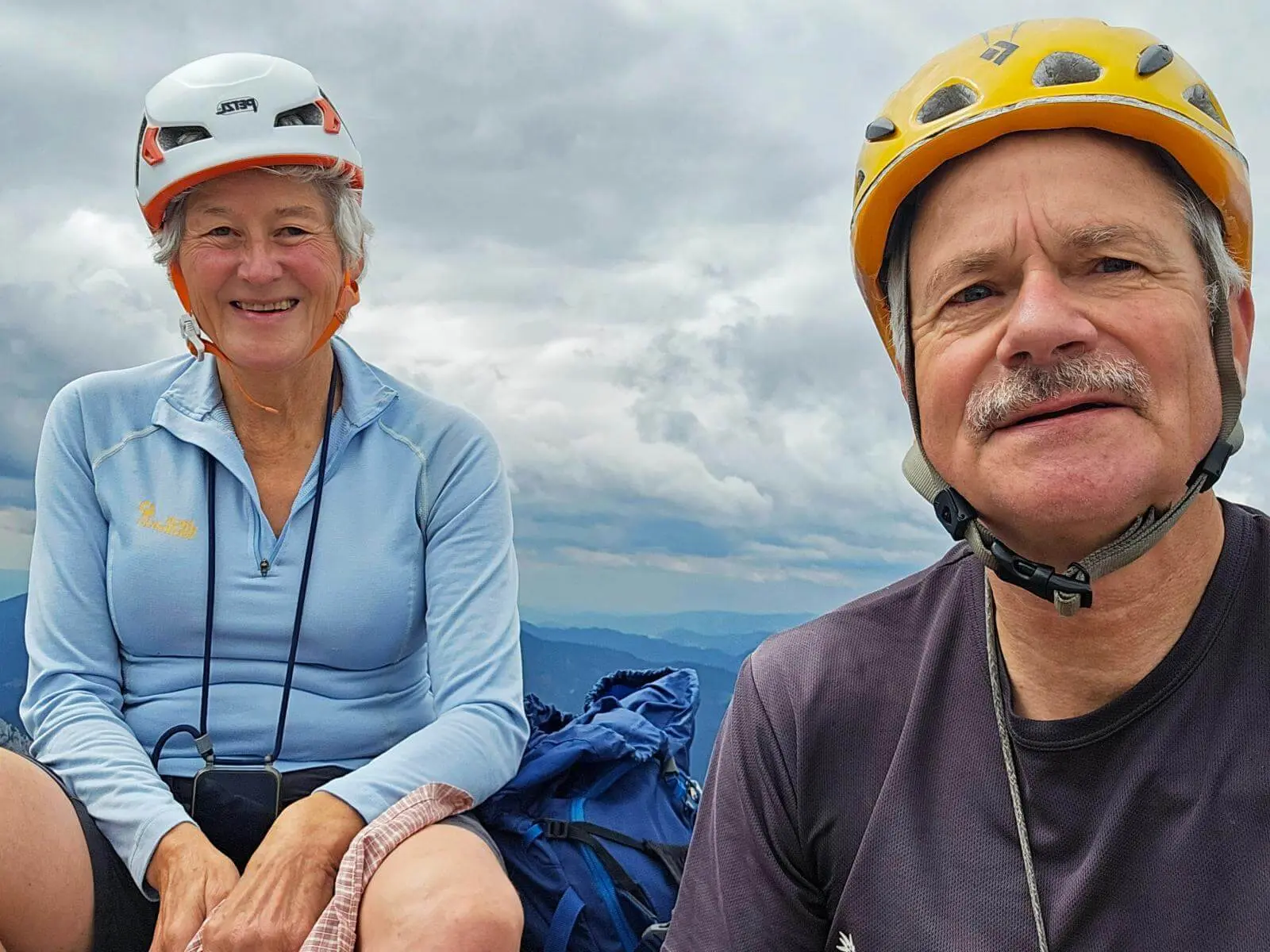 Zwei Personen mit Kletterhelmen sitzen auf einem Berggipfel vor wolkenverhangenem Himmel. Rucksäcke liegen neben ihnen am Boden. | © DAV Markt Schwaben/Lutz Gründel