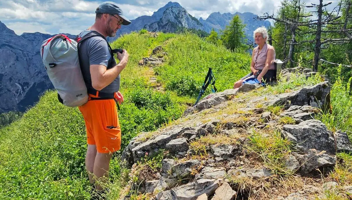 Eine Person mit Rucksack steht auf einem grasigen Bergrücken, während eine weitere auf Felsen sitzt. Dahinter erheben sich markante Gipfel unter wechselnder Bewölkung. | © DAV Markt Schwaben/Lutz Gründel