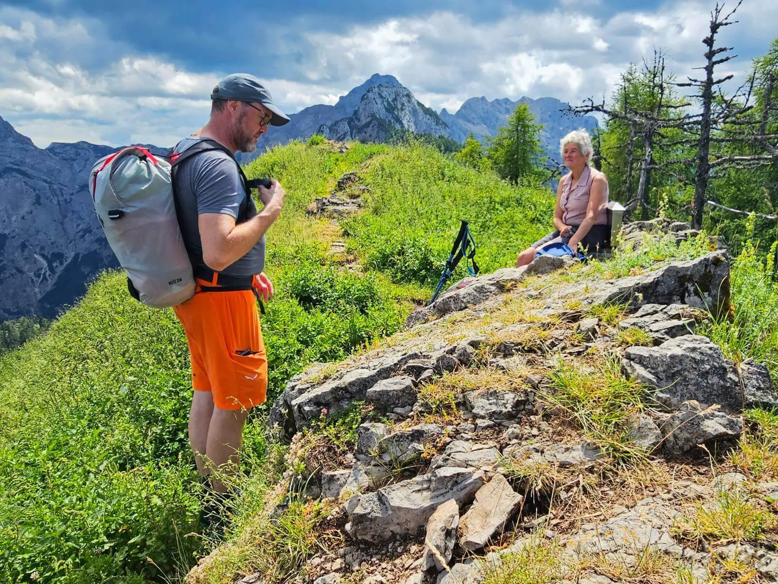 Eine Person mit Rucksack steht auf einem grasigen Bergrücken, während eine weitere auf Felsen sitzt. Dahinter erheben sich markante Gipfel unter wechselnder Bewölkung. | © DAV Markt Schwaben/Lutz Gründel