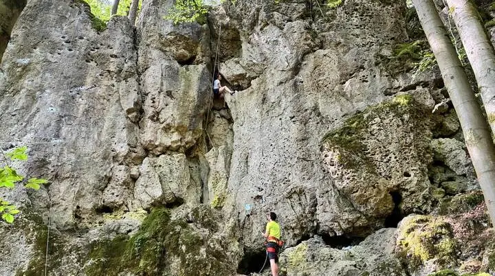 Eine Person klettert in einer steilen Felswand, während eine weitere Person am Boden sichert. Die zerklüfteten Kalkfelsen sind von Wald umgeben. | © DAV Markt Schwaben/Lara Wolff