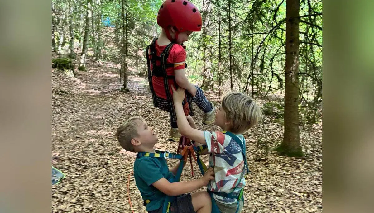 Drei Kinder mit Klettergurten und Helm üben im Wald den Umgang mit dem Seil. Ein Kind hängt gesichert am Seil, während die anderen unterstützen. | © DAV Markt Schwaben/Lara Wolff