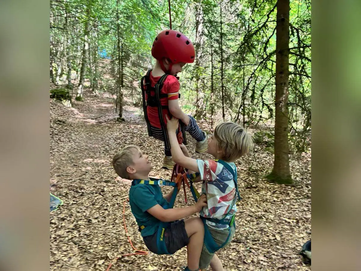 Drei Kinder mit Klettergurten und Helm üben im Wald den Umgang mit dem Seil. Ein Kind hängt gesichert am Seil, während die anderen unterstützen. | © DAV Markt Schwaben/Lara Wolff