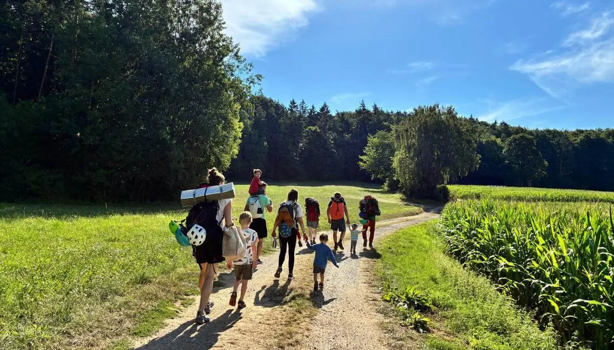 Mehrere Erwachsene und Kinder wandern mit Kletterausrüstung über einen Feldweg. Der Weg führt zwischen Wiesen und Waldrand zum Klettergebiet. | © DAV Markt Schwaben/Lara Wolff