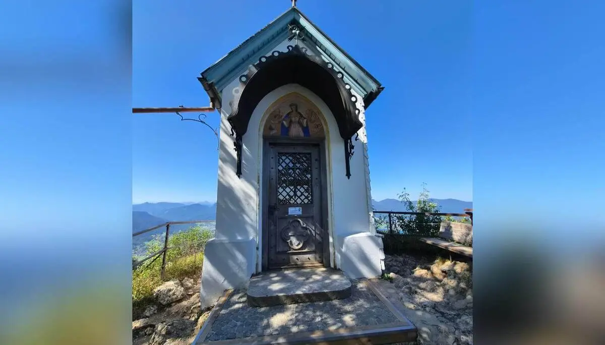 Kleine Kapelle auf dem Riederstein mit verziertem Eingang vor blauem Himmel; seitlich reicht der Blick in die Berglandschaft. | © DAV Markt Schwaben/Hubert Inhofer